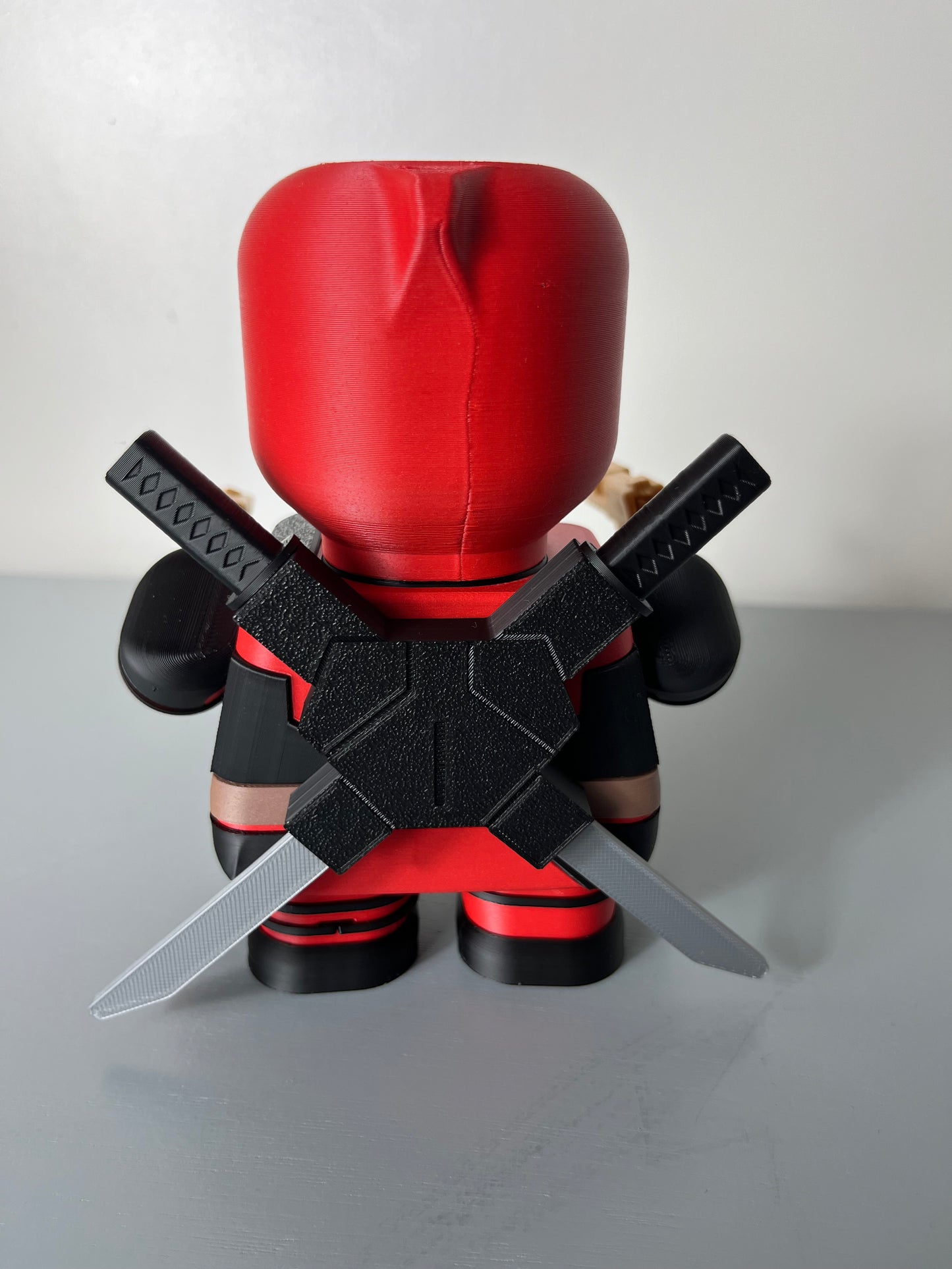 Back view of 3D printed superhero in red and black suit with mask, holding 2 guns and having 2 katana swords attached to his back