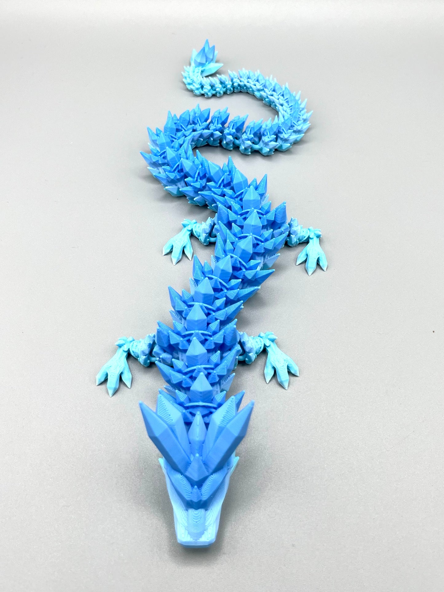 A 3D printed dragon in gradient shades of cool blue, featuring small detailed crystals all over, from head to tail. Set on a light grey background.