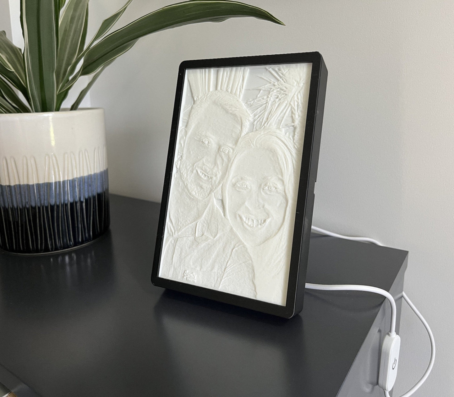 A custom 3D printed photo and frame displaying an image of two smiling faces, placed on a table with a plant in the background.