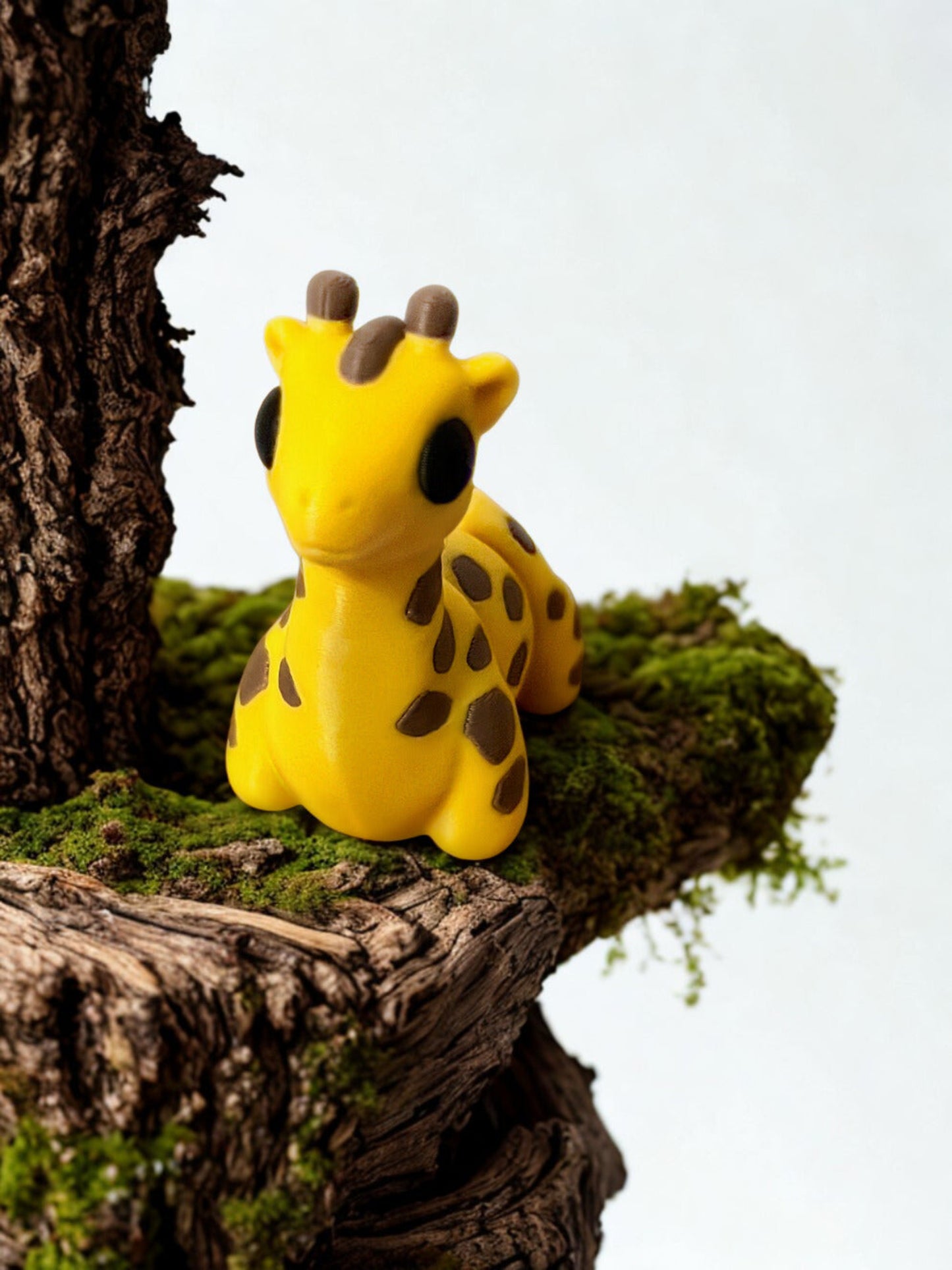 Small 3D printed yelloe giraffe figurine with brown spots on a mossy branch against a white background