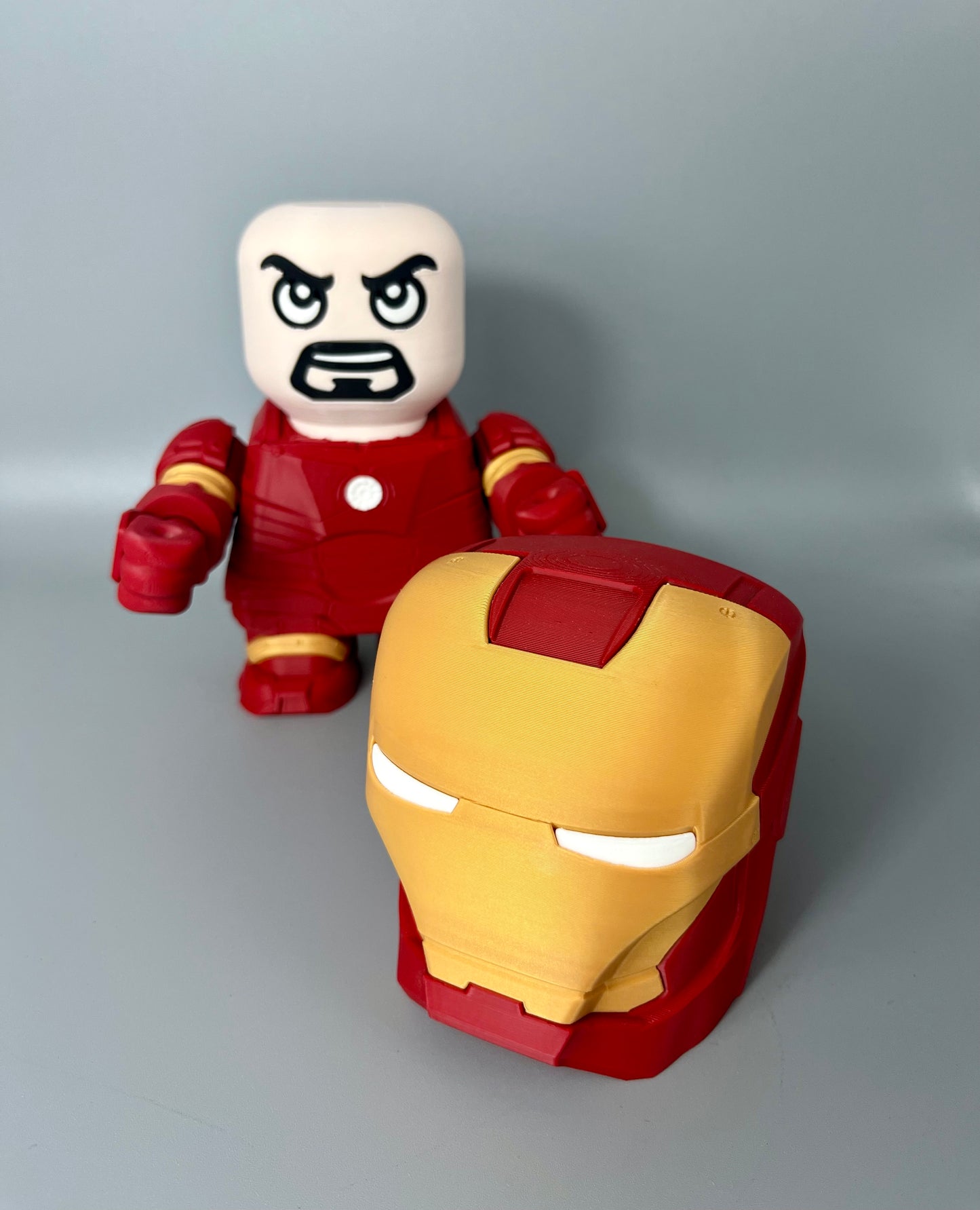 3D printed superhero with mask removed and on the floor, exposed face with goatee and red and gold superhero suit, set on a grey background