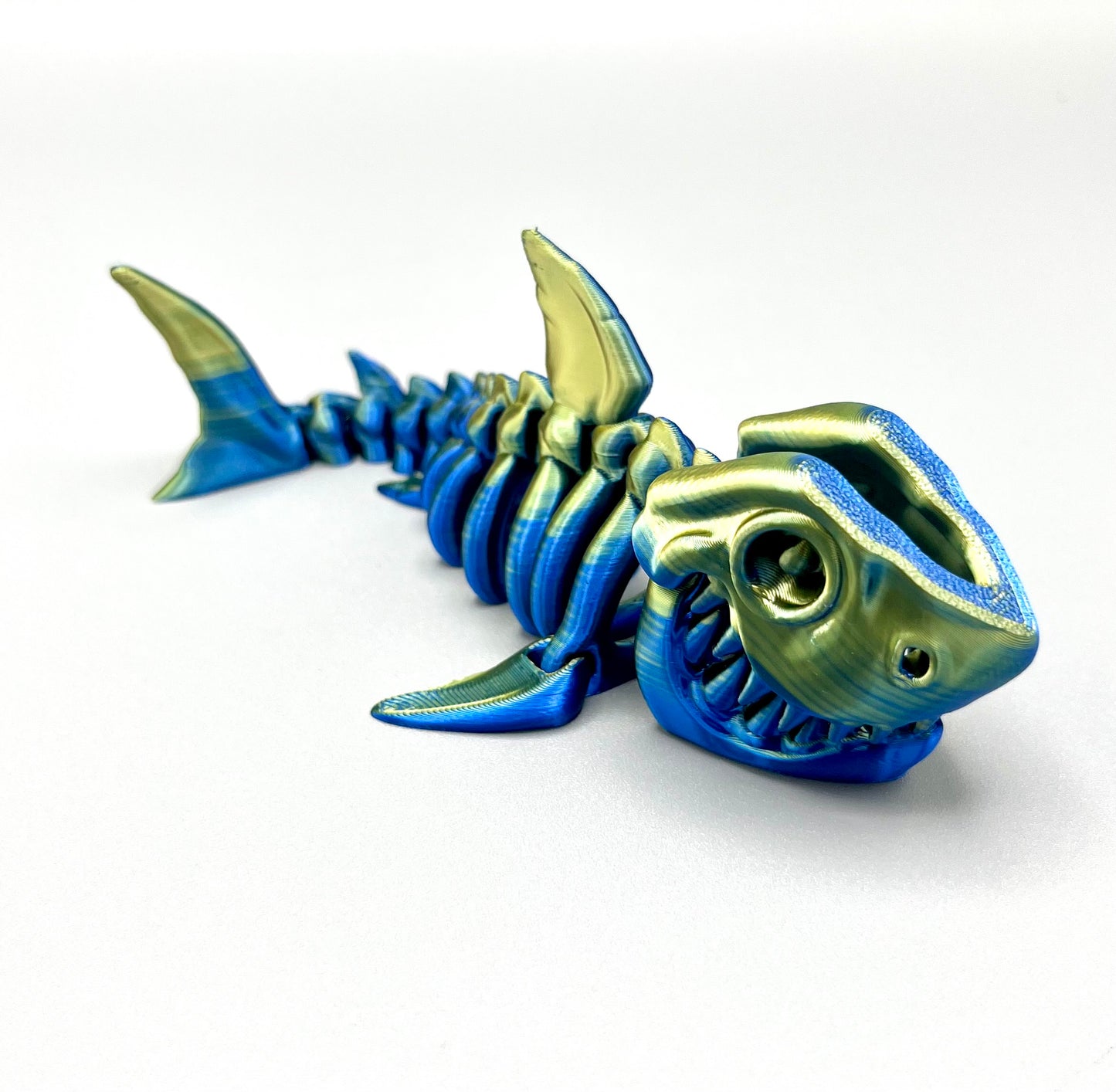 A small sized 3D printed gold and blue coloured shiny skeleton shark figurine with detailed features and wide mouth, showing teeth