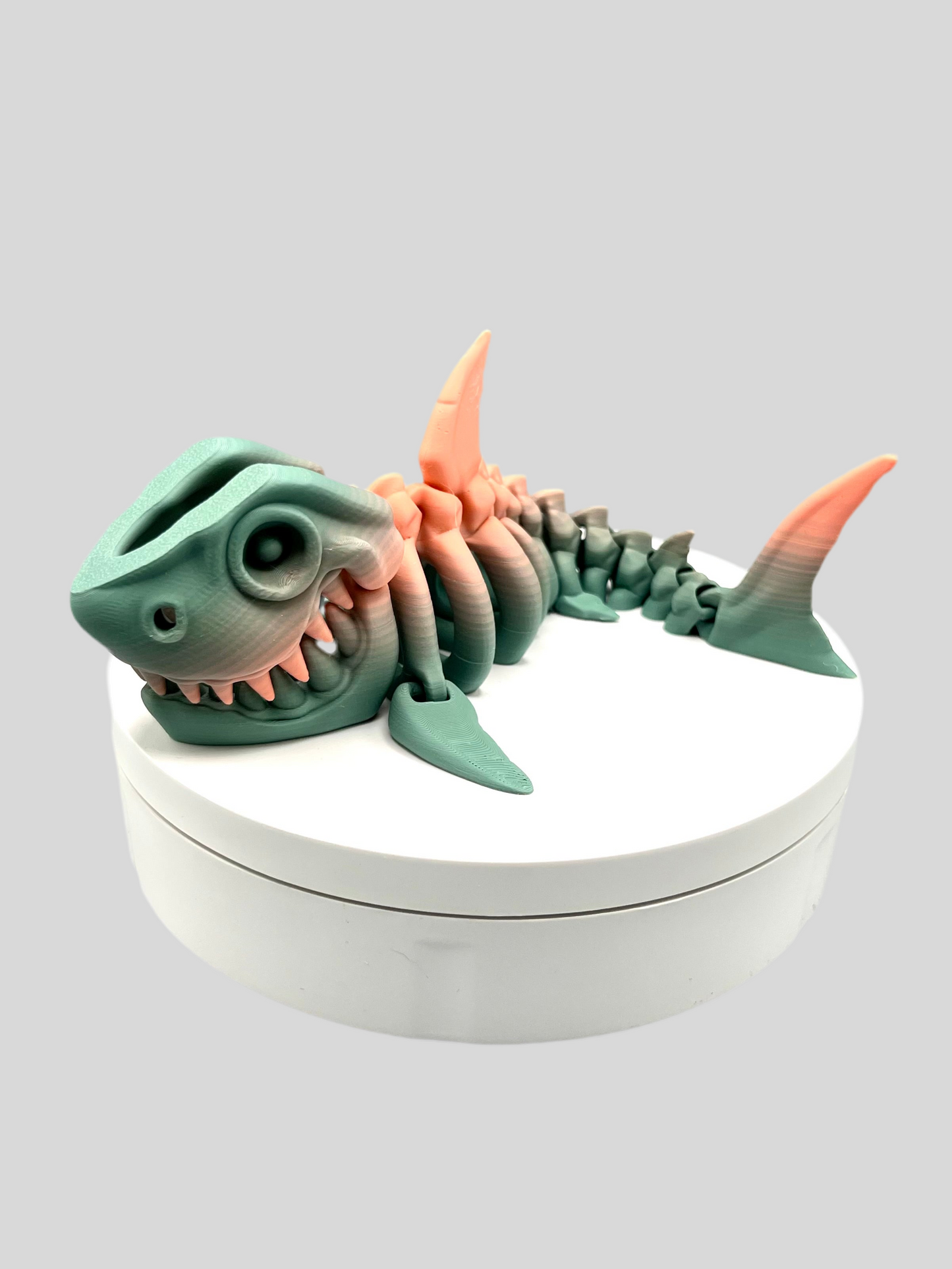 A small sized 3D printed khaki and coral coloured skeleton shark figurine with detailed features and wide mouth, showing teeth. Positioned on a white display base