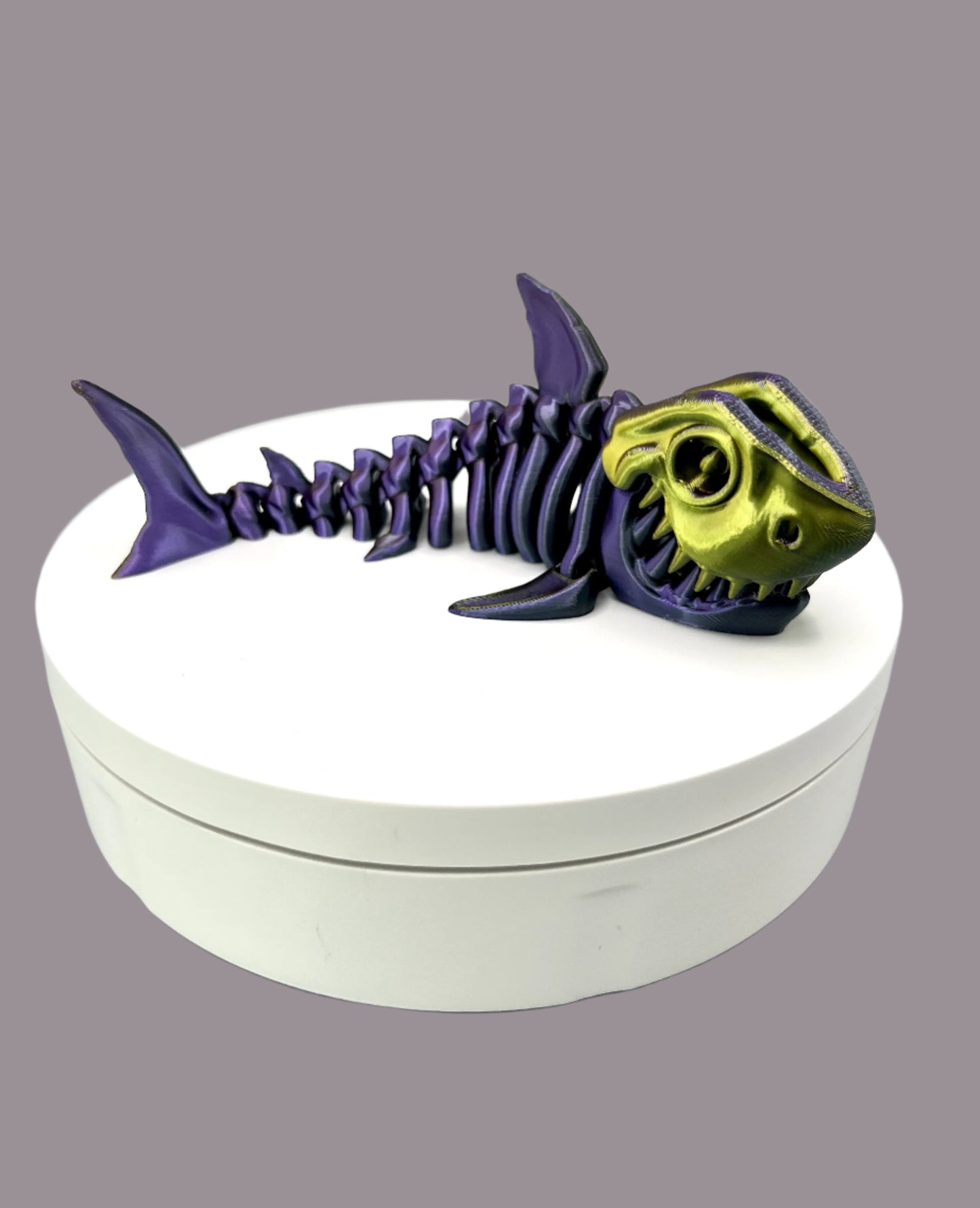A small sized 3D printed gold, purple and black coloured shiny skeleton shark figurine with detailed features and wide mouth, showing teeth. Positioned on a white display base