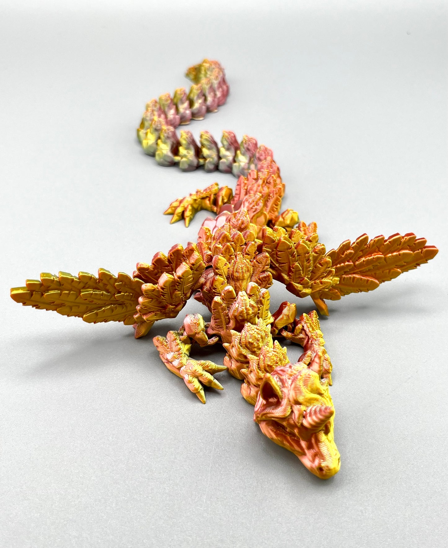 Metallic coloured winged 3D printed dragon with a unicorn's head and wings outstretched. Set on a light grey background.