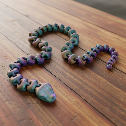 An articulated 3D-printed snake, in a shimmery finish and featuring realistic facial features. Laying on a wooden table.