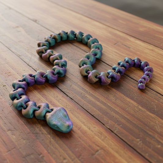 An articulated 3D-printed snake, in a shimmery finish and featuring realistic facial features. Laying on a wooden table.