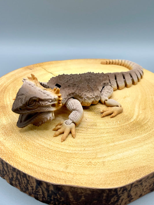 A 3D printed coffee gradient coloured Bearded Dragon, with realistic features, textured face and body and articulated tail. Sitting on wood. 