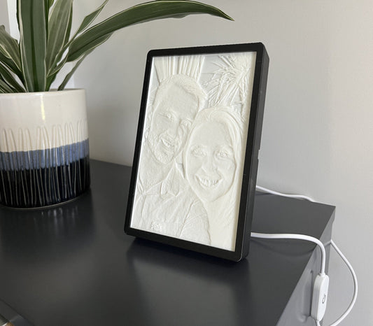 A custom 3D printed photo and frame displaying an image of two smiling faces, placed on a table with a plant in the background.