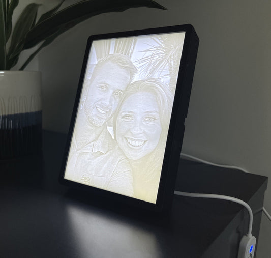A custom 3D printed photo and frame displaying an image of two smiling faces, placed on a table with a plant in the background.