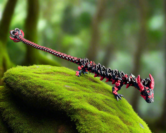 A long, highly detailed 3D printed dragon with black and red love hearts printed along its articulated body, positioned standing on a mossy rock in a rainforest