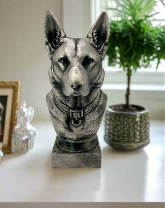 A black and silver 3D printed bust of a German Shepherd displayed on a white table in front of a window and next to a plant in a green ceramic pot 