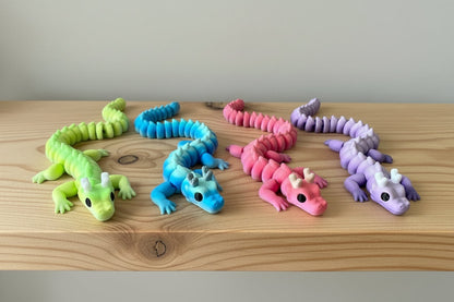 Four 3D printed articulated dragons with horns, crafted in vibrant PLA filament, featuring flexible segmented bodies; suitable for fidgeting or display. Sat on a wooden shelf against a white wall. 