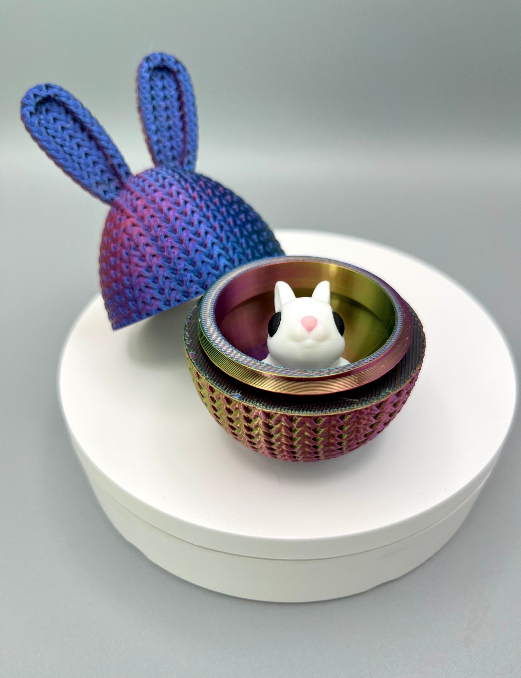 A 3D printed knitted bunny ear egg, opened up to show a little bunny inside. Displayed on a round white surface.