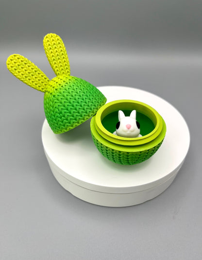 A 3D printed knitted bunny ear egg, opened up to show a little bunny inside. Displayed on a round white surface.