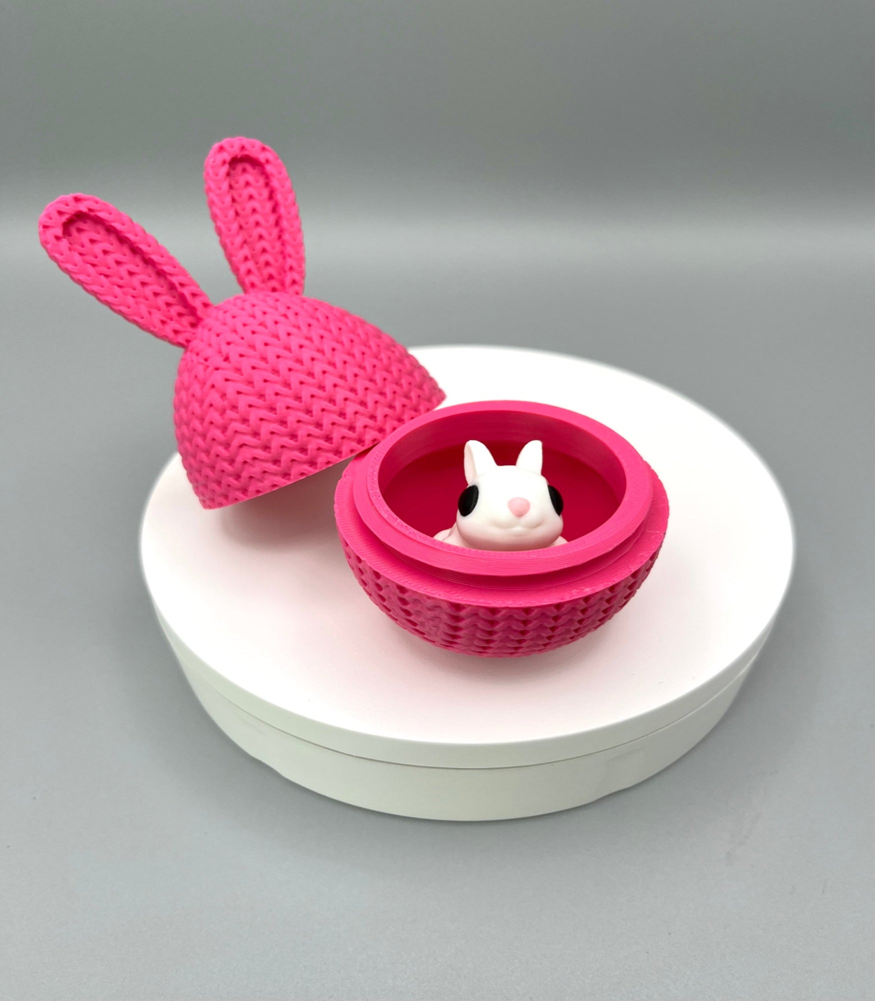 A 3D printed knitted bunny ear egg, opened up to show a little bunny inside. Displayed on a round white surface.