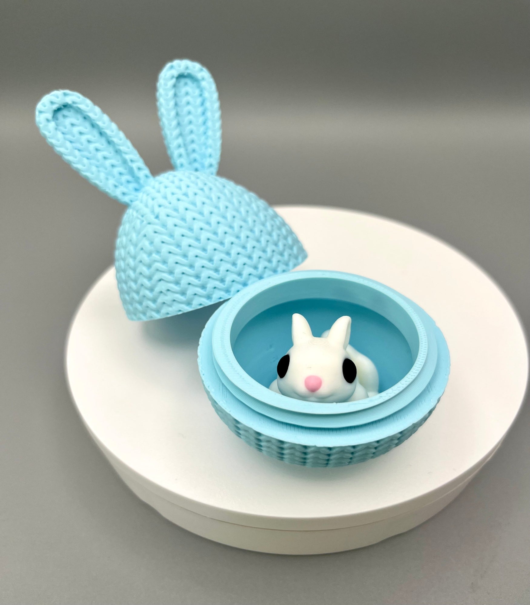 A 3D printed knitted bunny ear egg, opened up to show a little bunny inside. Displayed on a round white surface.