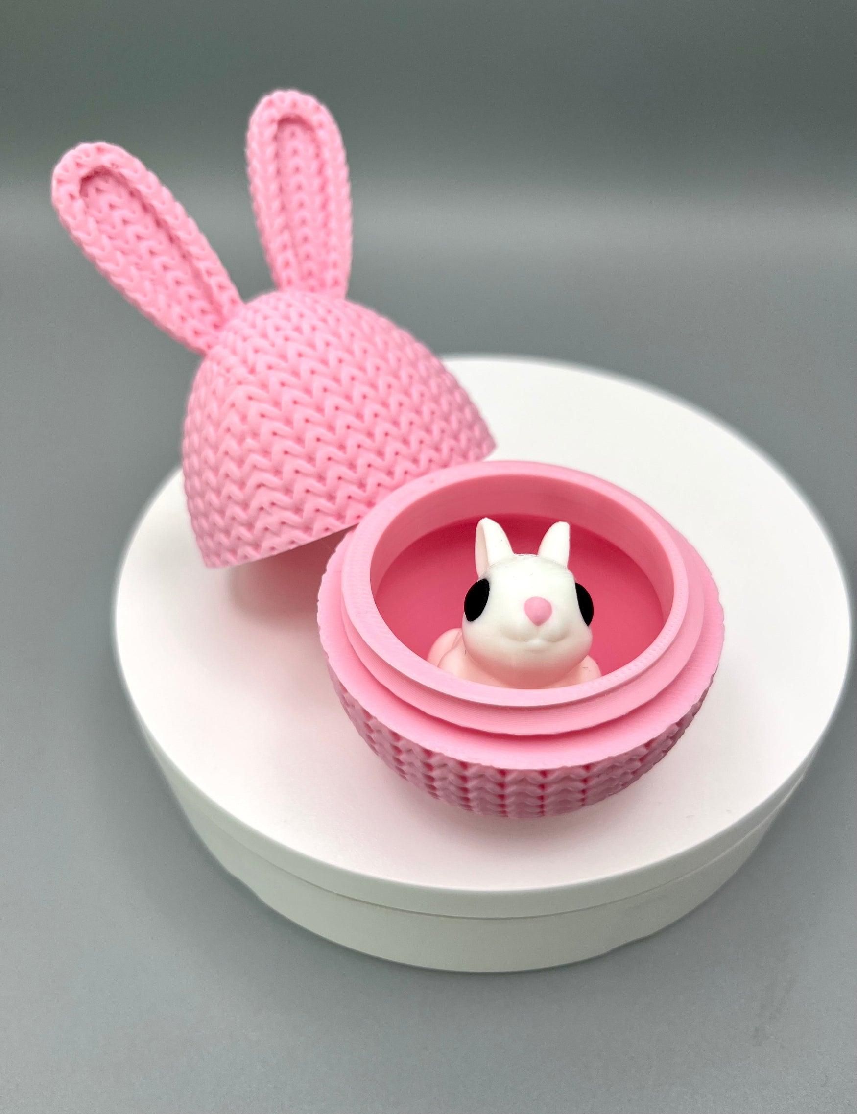 A 3D printed knitted bunny ear egg, opened up to show a little bunny inside. Displayed on a round white surface.