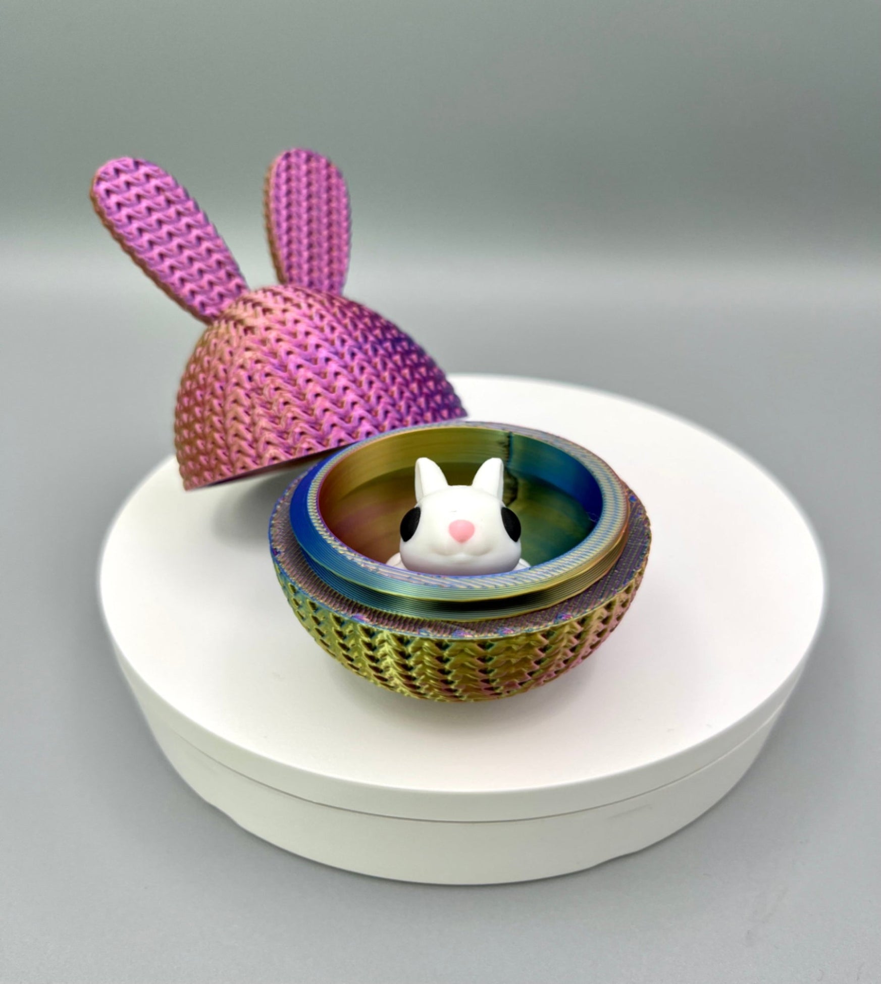 A 3D printed knitted bunny ear egg, opened up to show a little bunny inside. Displayed on a round white surface.