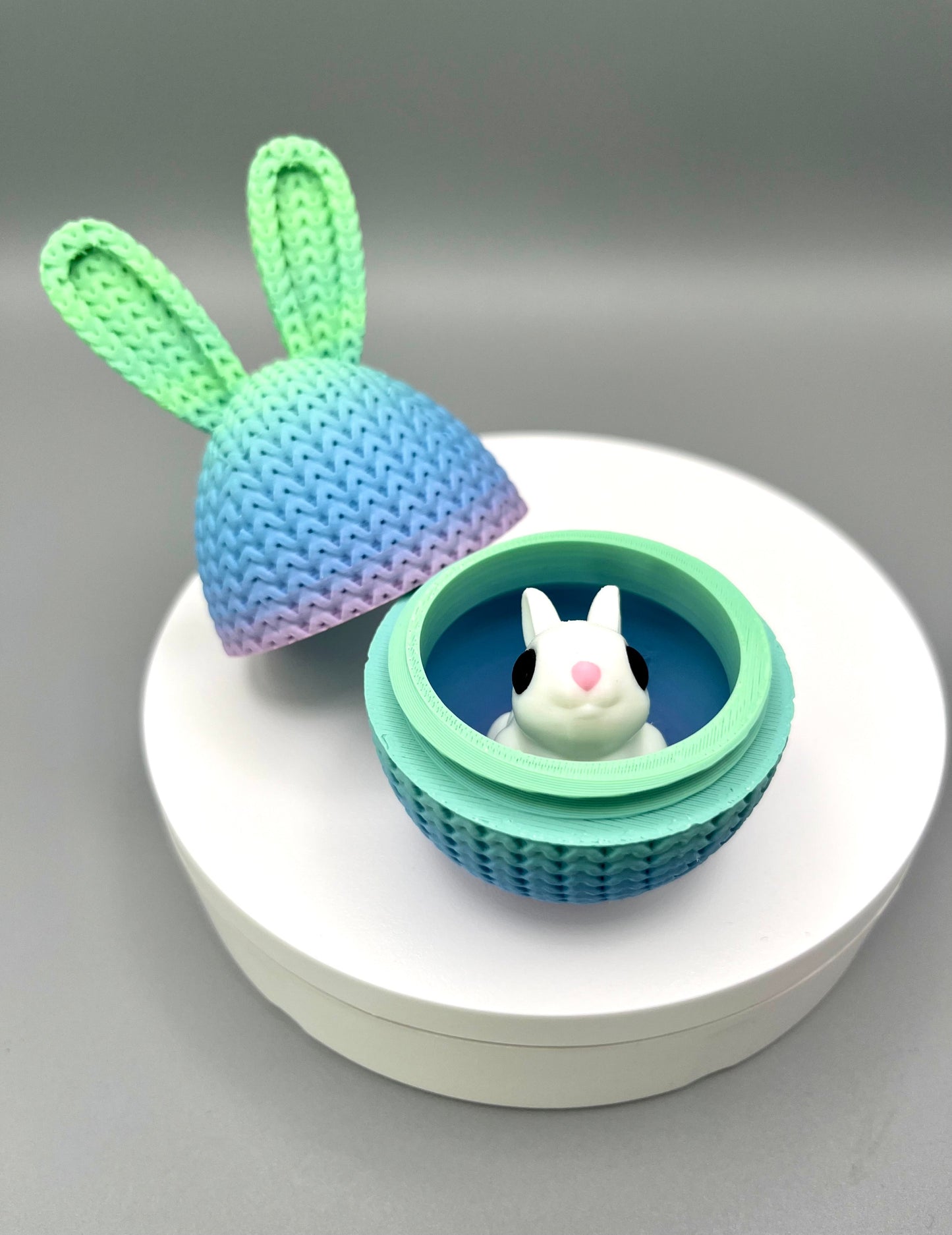 A 3D printed knitted bunny ear egg, opened up to show a little bunny inside. Displayed on a round white surface.