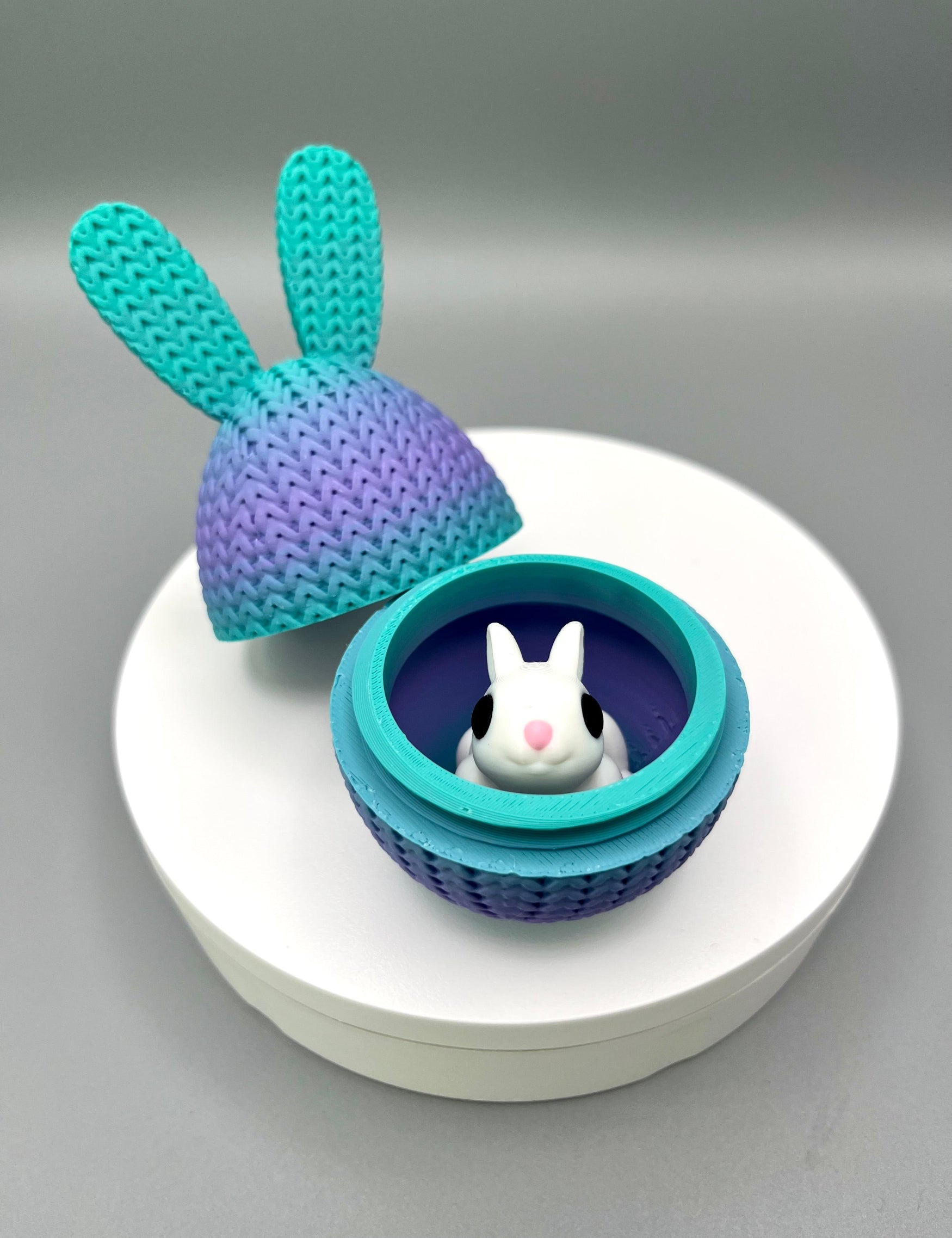 A 3D printed knitted bunny ear egg, opened up to show a little bunny inside. Displayed on a round white surface.