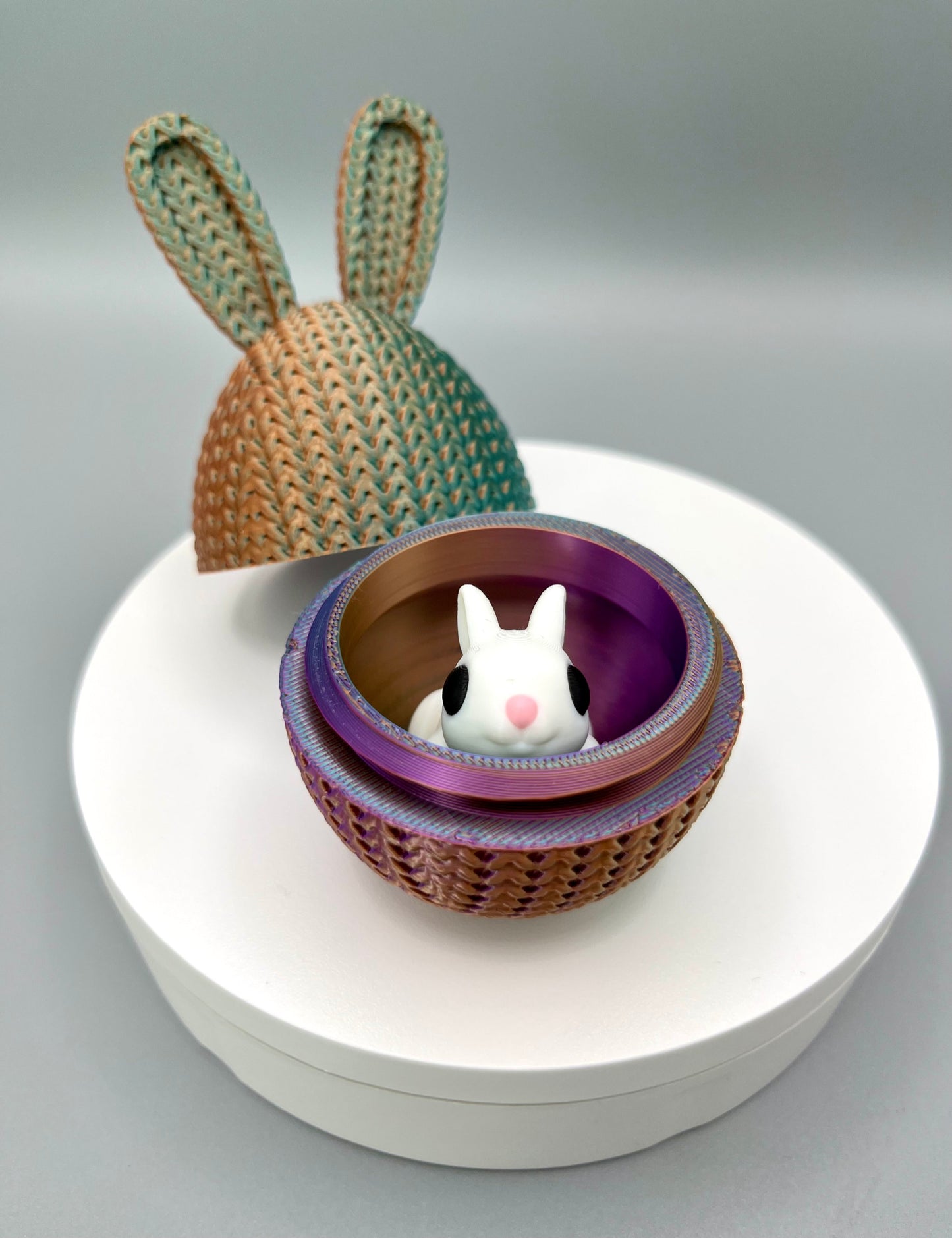 A 3D printed knitted bunny ear egg, opened up to show a little bunny inside. Displayed on a round white surface.