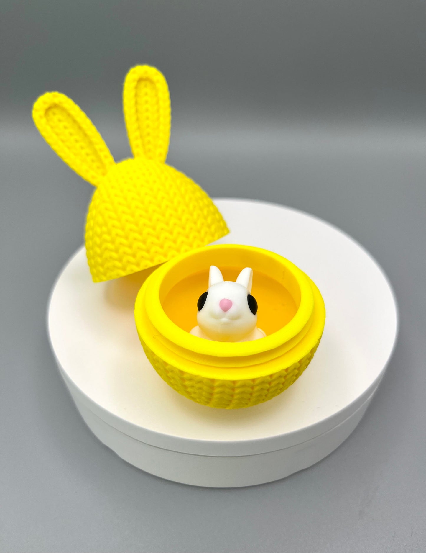 A 3D printed knitted bunny ear egg, opened up to show a little bunny inside. Displayed on a round white surface.