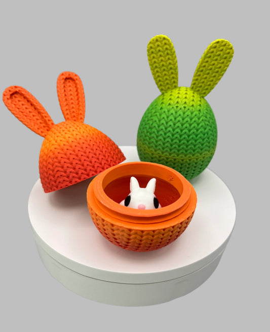 A 3D printed collection of 2 knitted bunny ear eggs in orange and green; one is opened and has a white bunny inside. Both are displayed on a round white surface.