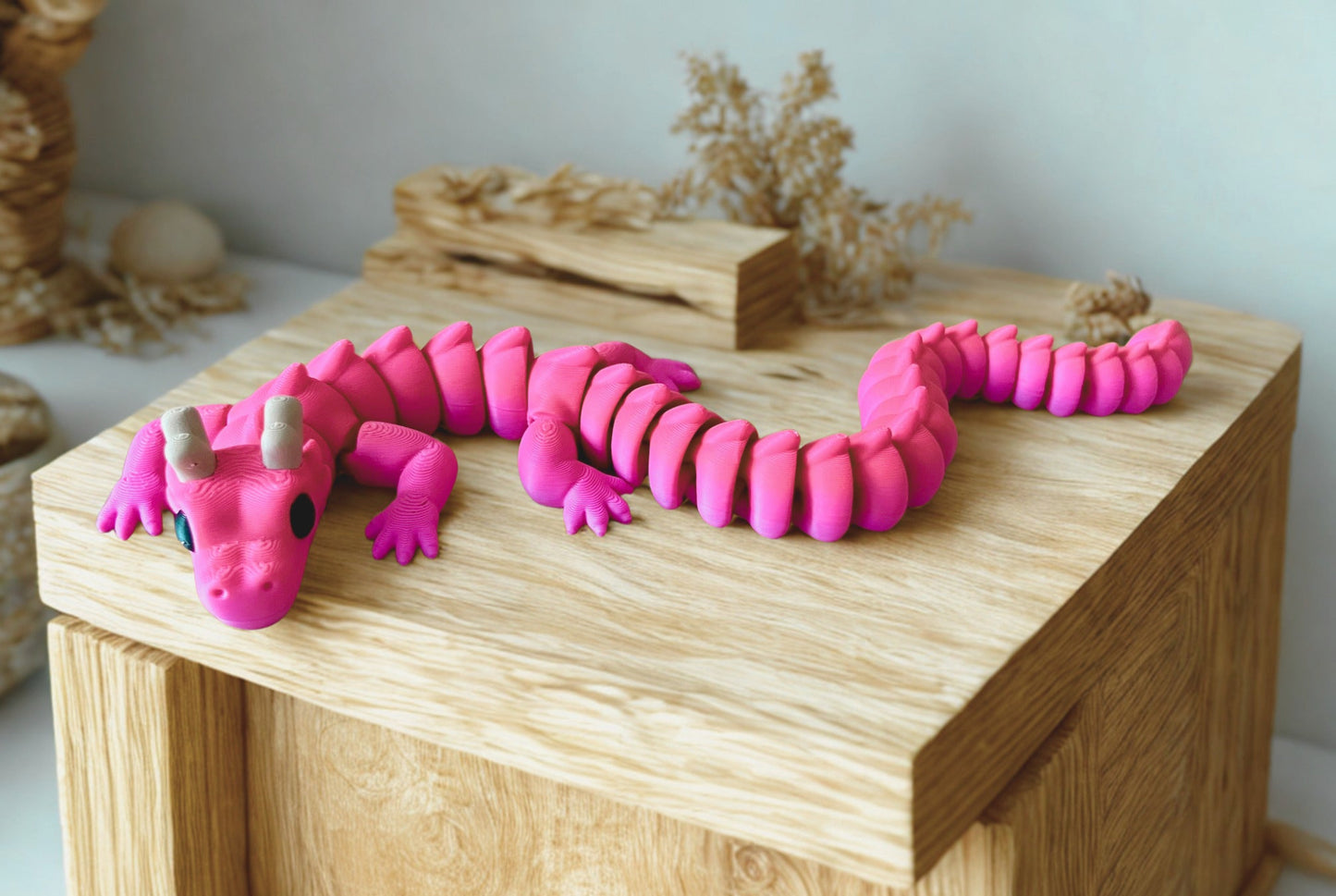 Colourful 3D printed articulated dragon with horns, lying on a wooden shelf . It has a flexible, segmented body and moveable little legs.