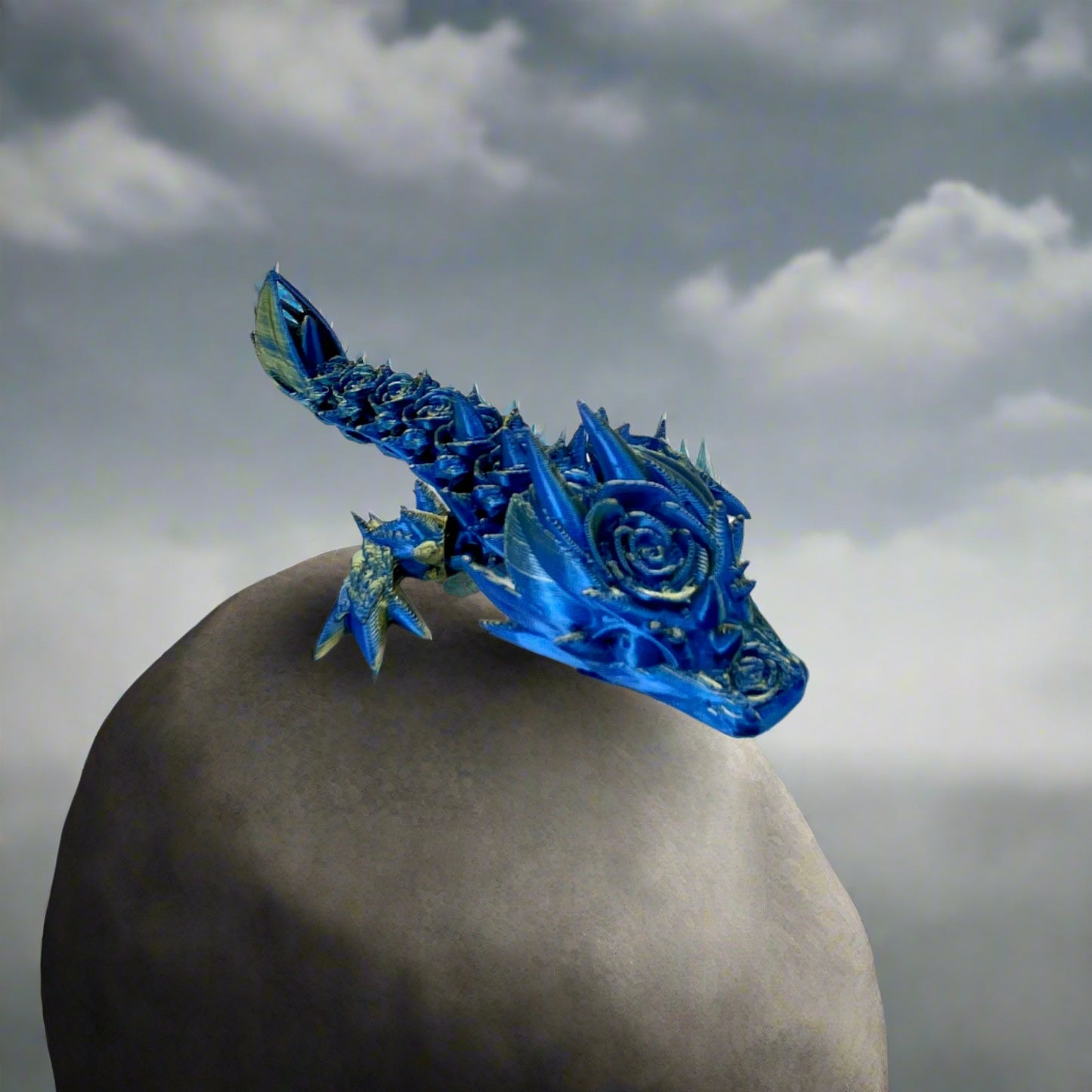 A small 3D-printed dragon with a prominent rose design on its head and small roses trailing along its body. Sat on a large grey rock