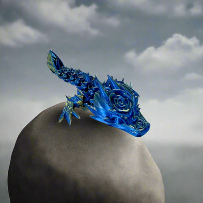 A small 3D-printed dragon with a prominent rose design on its head and small roses trailing along its body. Sat on a large grey rock