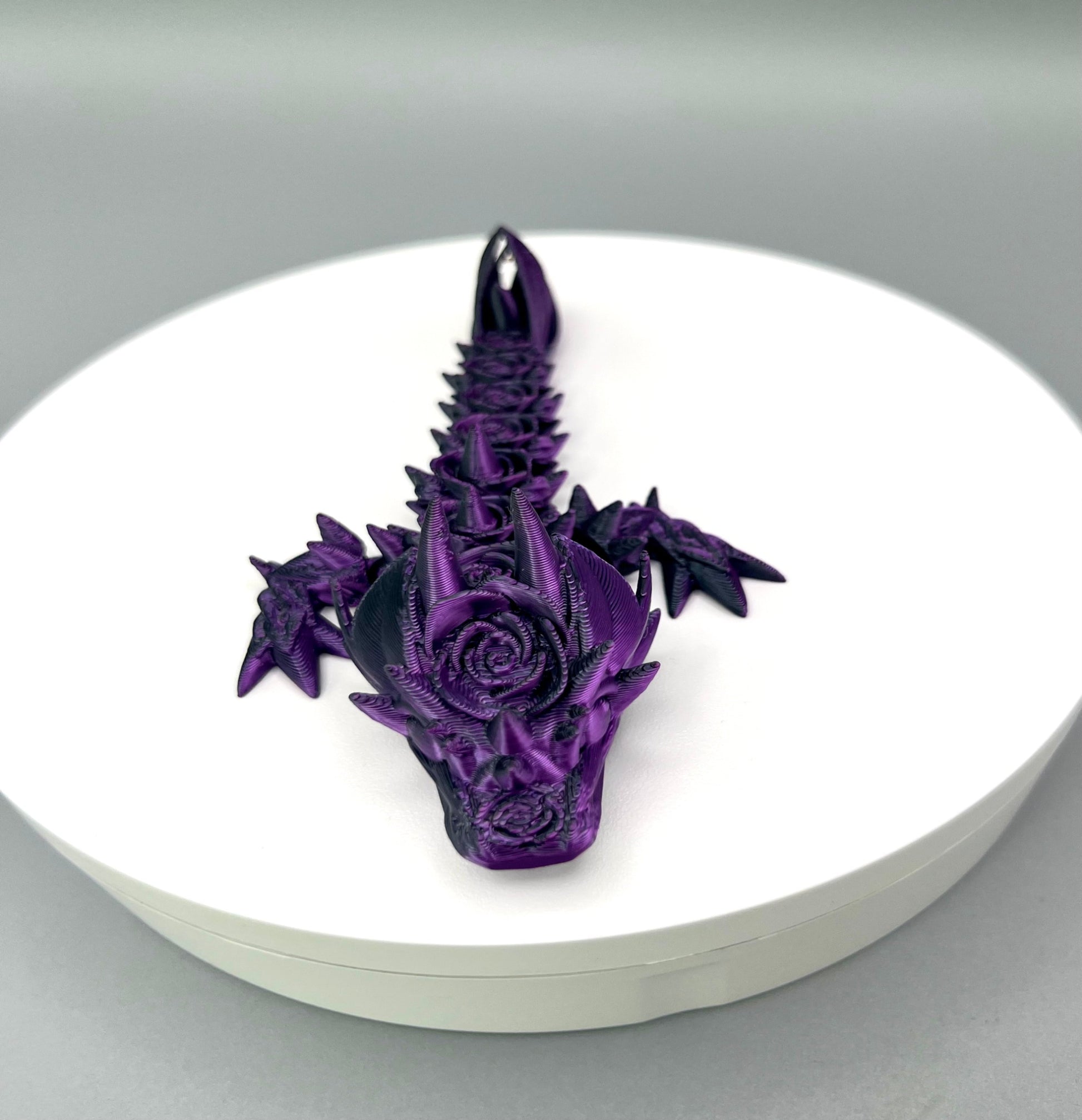 A small 3D-printed dragon with a prominent rose design on its head and small roses trailing along its body. Sat on a white circular base.