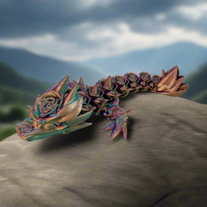 A small 3D-printed dragon with a prominent rose design on its head and small roses trailing along its body. Sat on a rock with countryside in the background