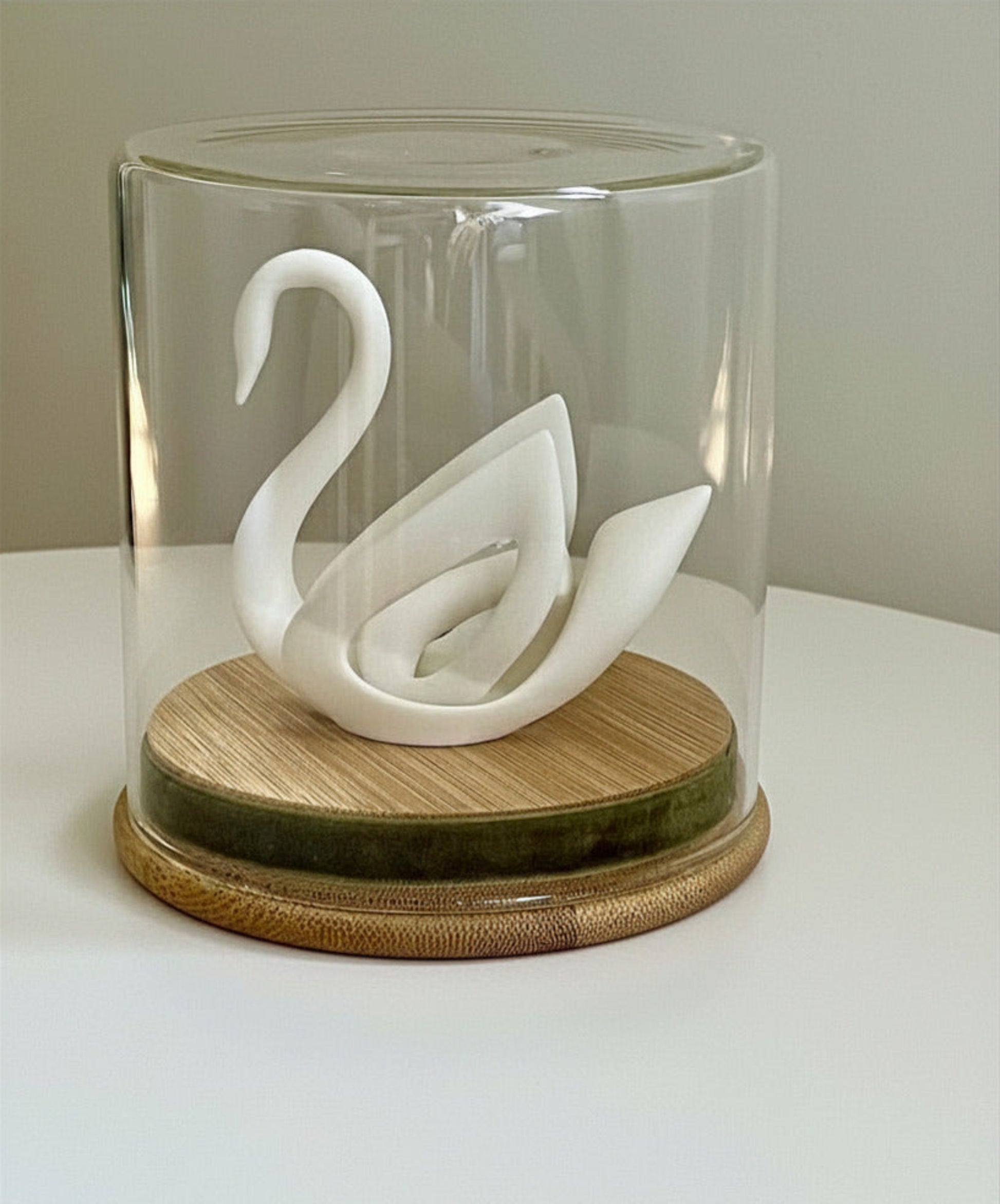 A white 3D printed swan displayed under a clear glass dome on a wooden base wrapped with a moss‑green ribbon. Positioned on a white surface. 