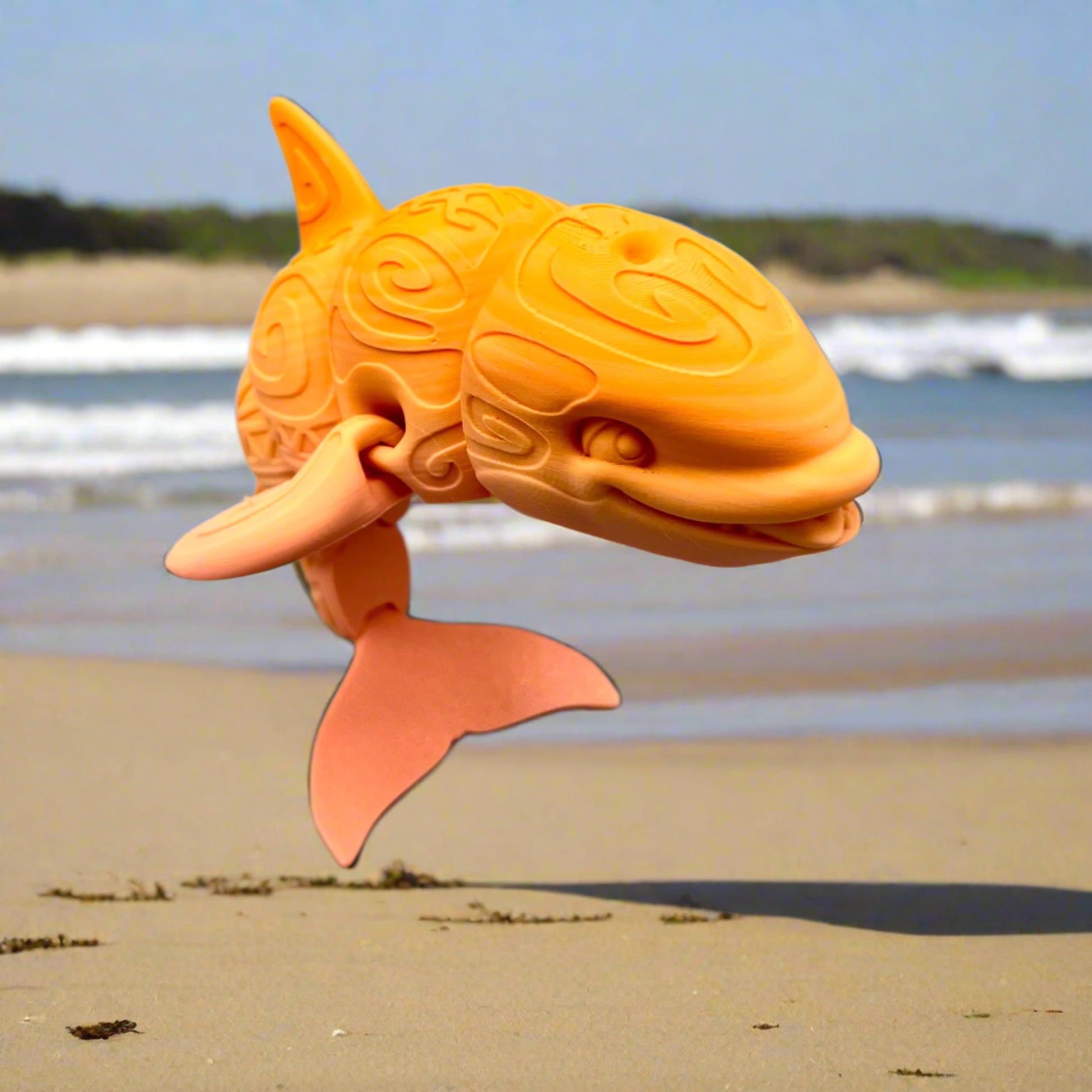 A 3D-printed orca with a tattooed all-over print, detailed facial features, including a smile, exposed teeth and tongue. Positioned sanding on it's tail on a sandy beach with rolling waves coming in. 