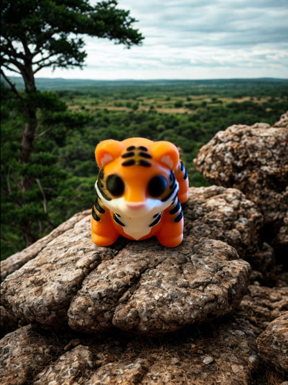 Small 3D‑printed tiger figurine with orange and black stripes, sitting upright on a rocky surface with a natural outdoor background.